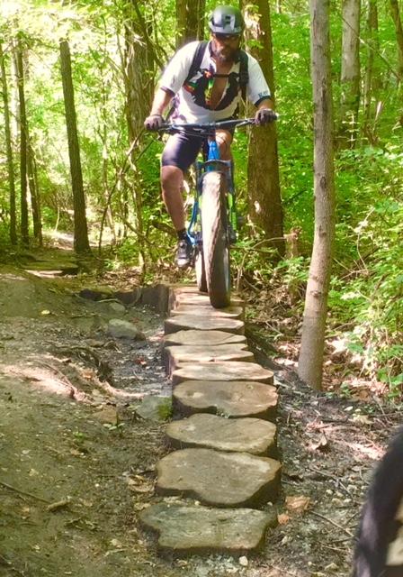 A person riding a mountain bike on a narrow stone path through a lush green forest. The cyclist is wearing a helmet and a backpack, navigating over natural stone stepping stones surrounded by trees and greenery. Perimeter Trail mountain bike trail.