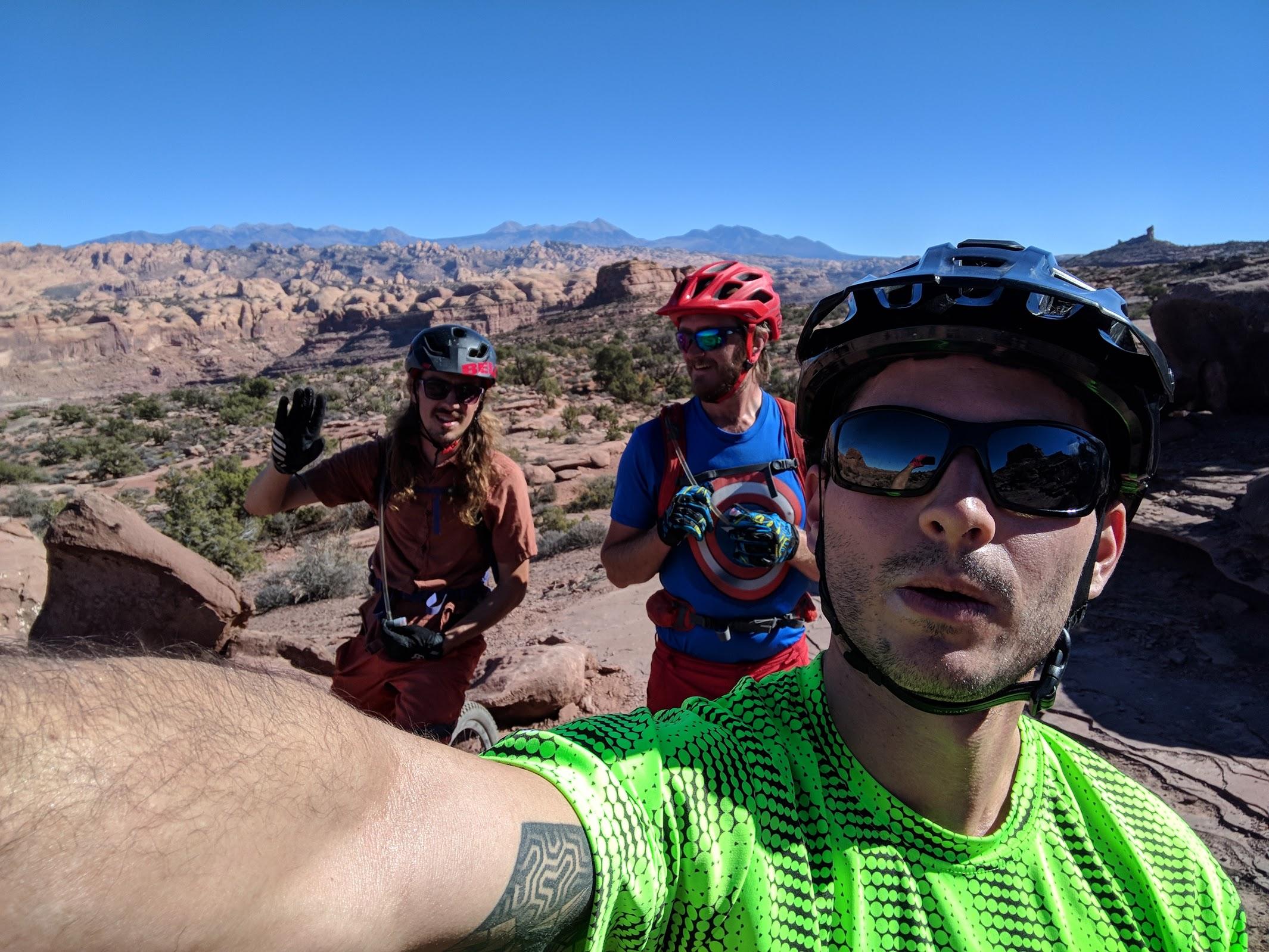 Three mountain bikers pose for a selfie in a rocky outdoor setting, with mountains in the background under a clear blue sky. One biker, wearing a bright green shirt, is in the foreground, while the other two, in helmets and cycling gear, are slightly behind, with one waving. The scene captures the adventure and camaraderie of biking in nature. Captain Ahab mountain bike trail.