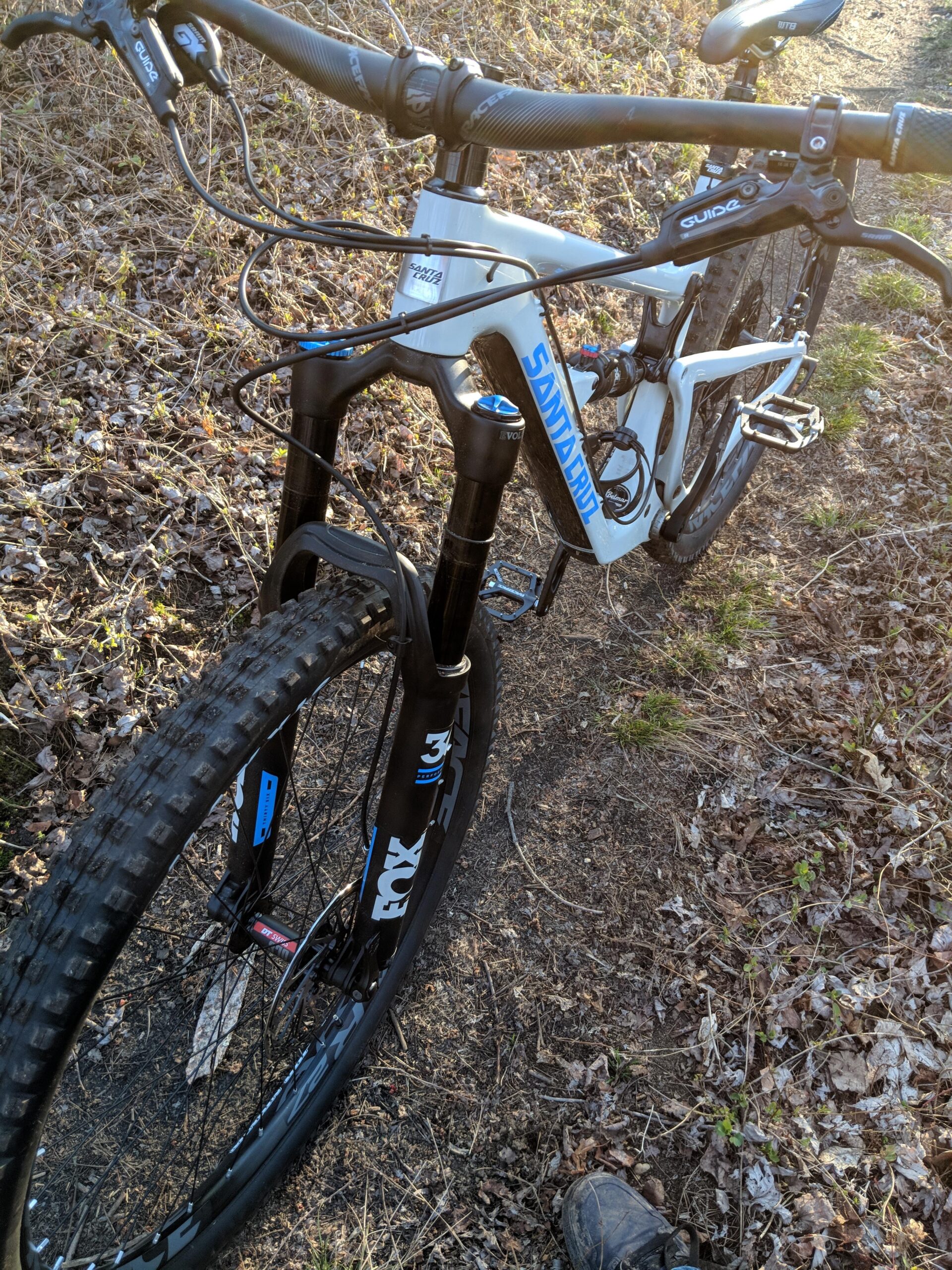 Santa Cruz Hightower 29er/27.5: A close-up view of a mountain bike on a dirt trail, featuring a white and blue frame with the brand name "Santa Cruz." The handlebars, front fork, and tire are clearly visible, showcasing the bike's suspension and rugged design. The background includes earthy textures with scattered leaves, indicating a natural outdoor setting.