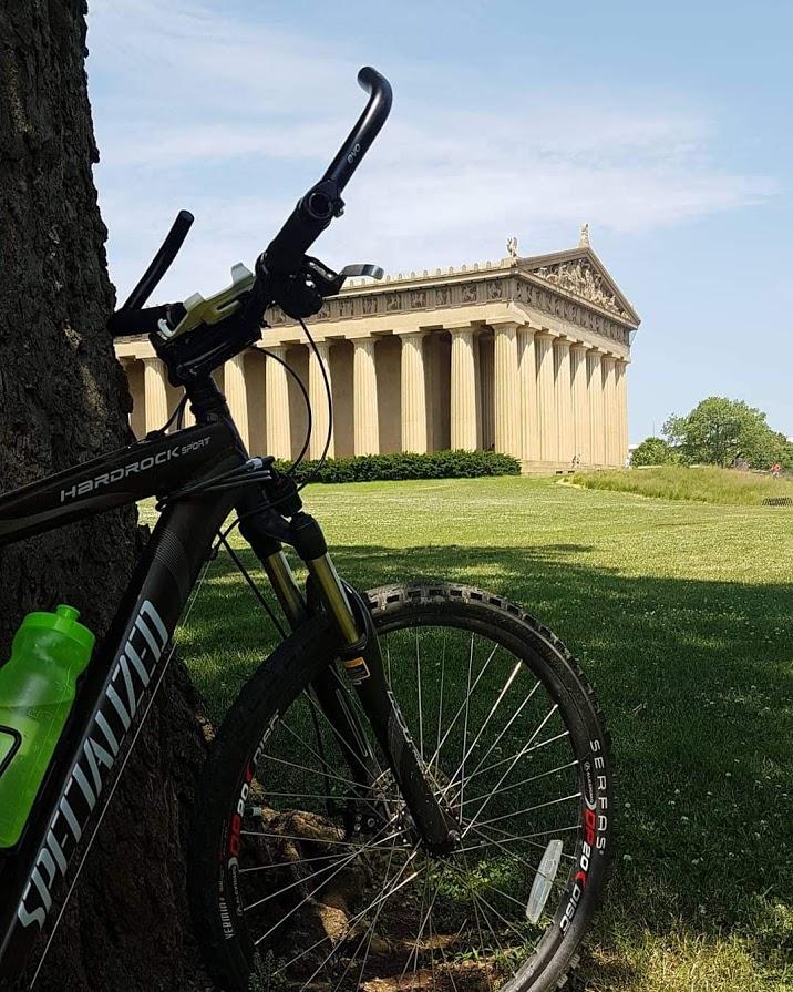 Specialized Hardrock Sport Disc: A mountain bike is leaning against a tree with a water bottle attached, framed by a grassy area. In the background, a large classical-style building with columns and a pediment is visible under a clear blue sky.