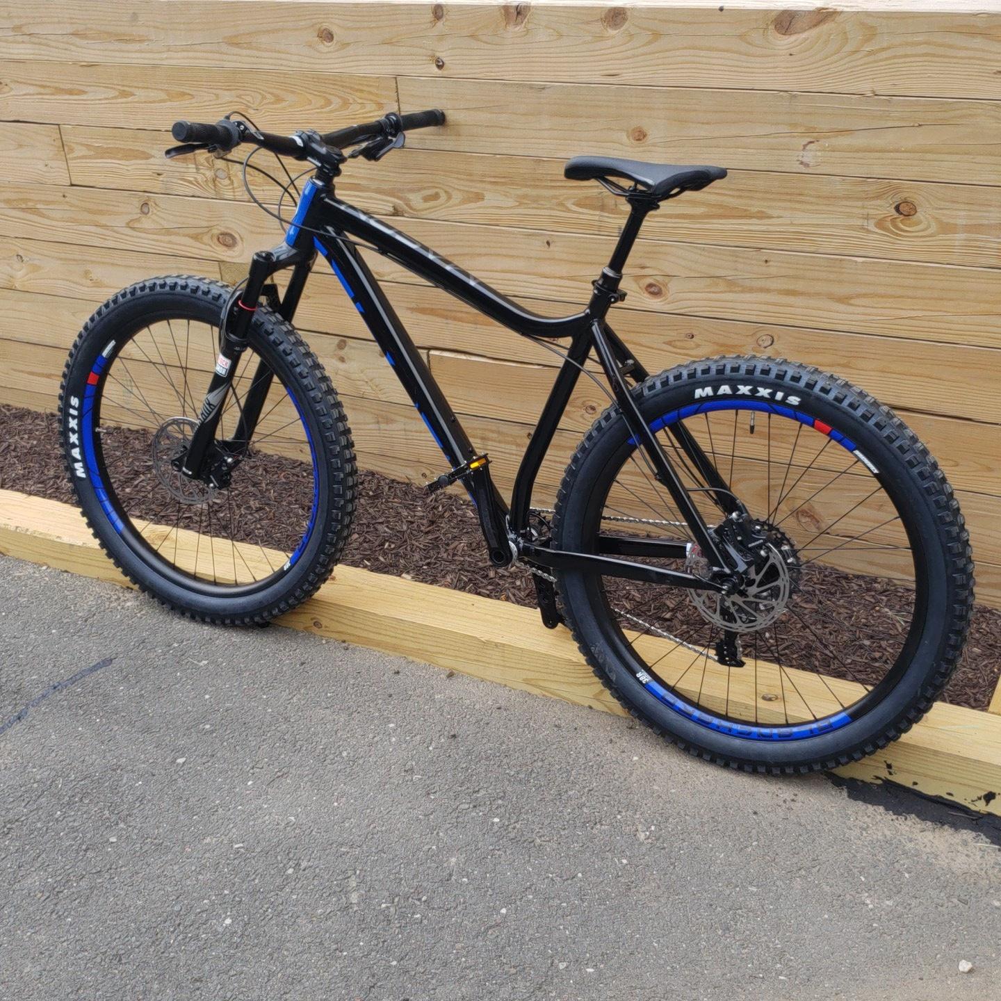 Diamondback Mason 2: A black mountain bike with blue accents, positioned against a wooden wall. The bike features thick, knobby tires labeled "MAXXIS," a lightweight frame, and a simple design suitable for off-road cycling.