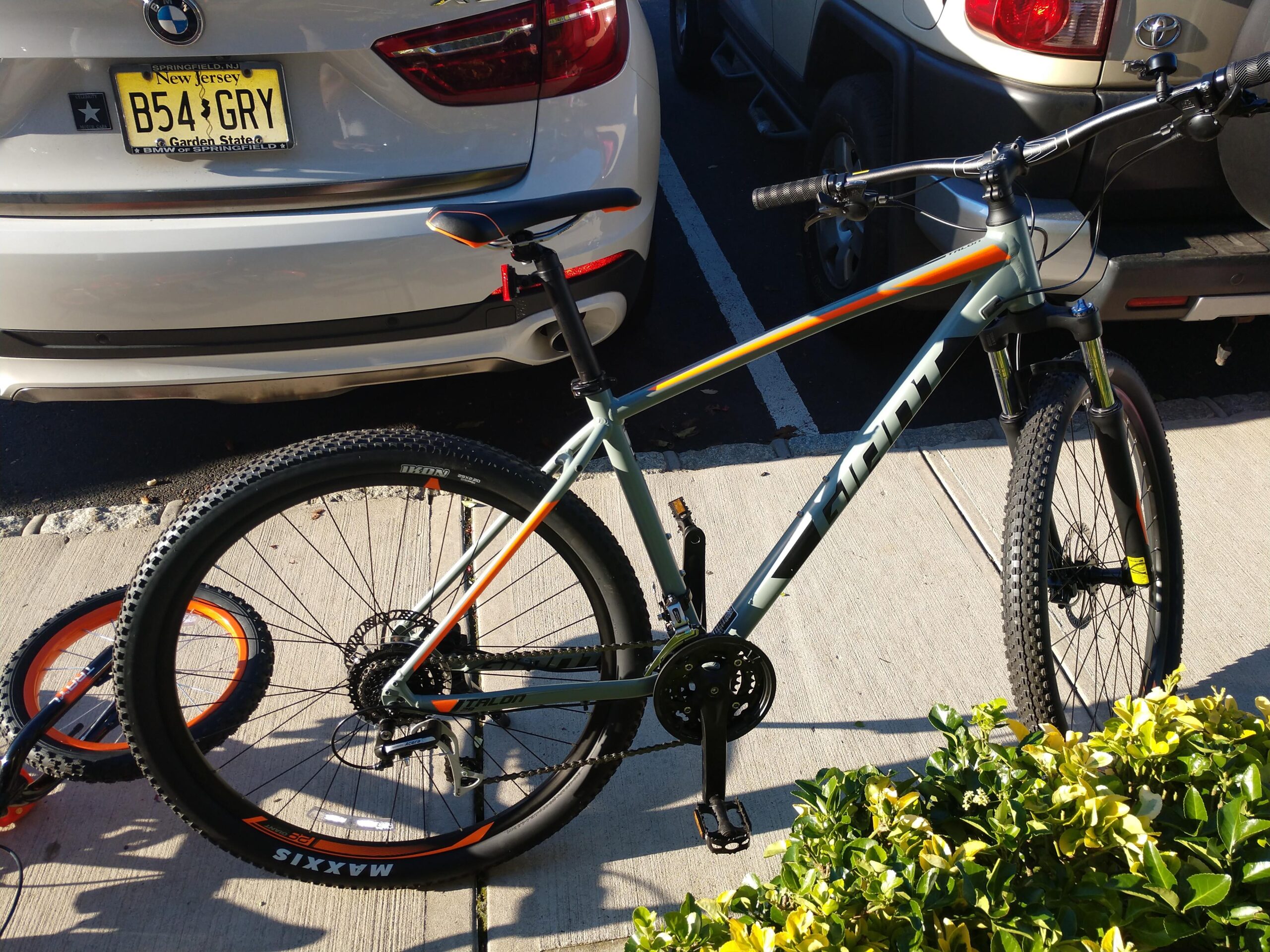 Giant Talon 29er: A mountain bike is parked next to a gray car in a parking lot, with a partially visible tire resting on the ground. The bike has a light green frame with orange and gray accents, and it features thick, knobby tires. Surrounding the bike are some green shrubs and the pavement of the parking area.