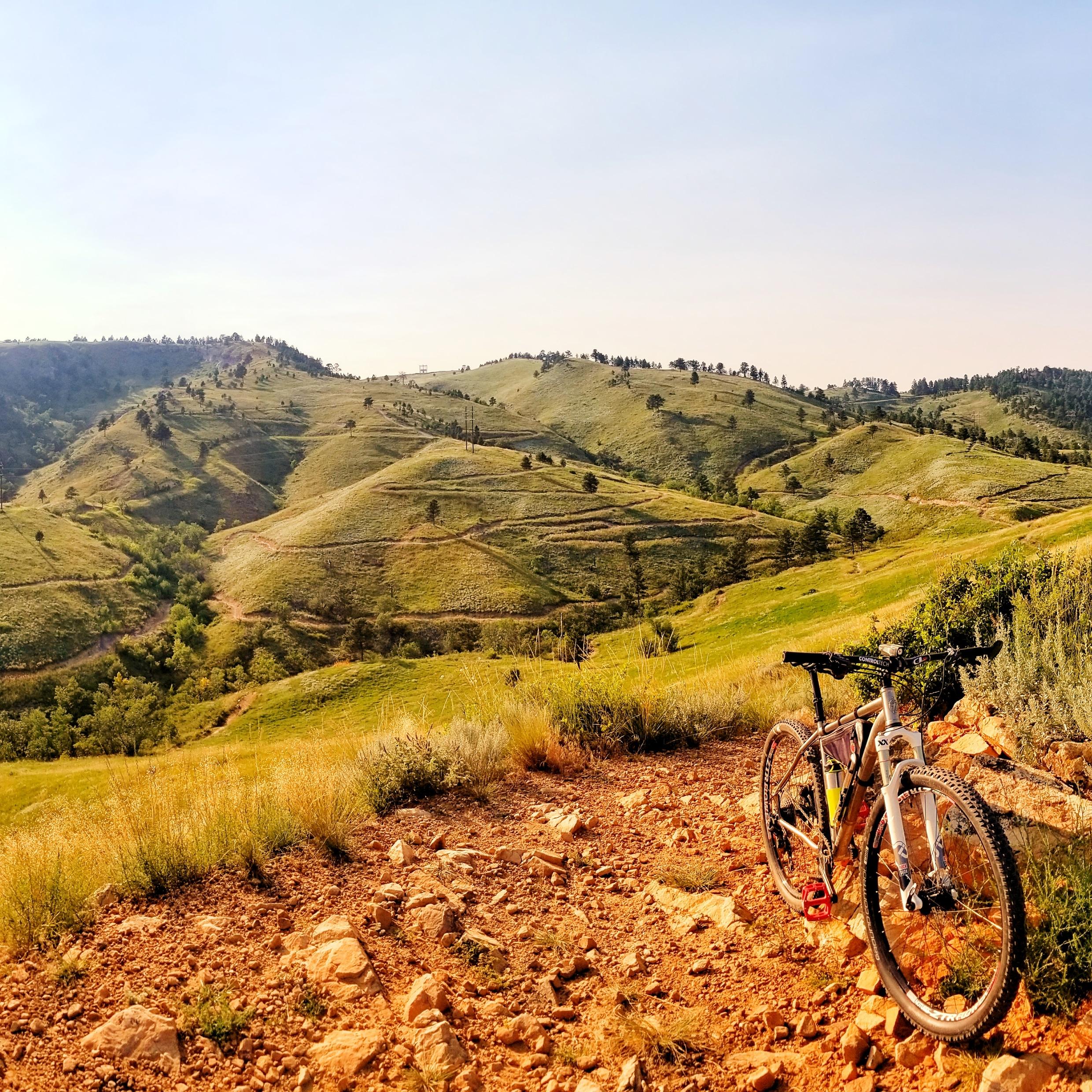 Mountain bike resting on a gravel path with rolling green hills and trees in the background under a clear sky. HLMP mountain bike trail.
