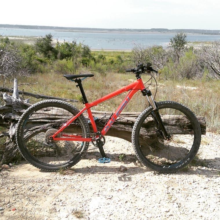 A bright orange mountain bike is parked beside a fallen log in a natural landscape, with a lake and distant hills in the background. The surroundings feature green grass and shrubs under a clear sky. Dana Peak mountain bike trail.