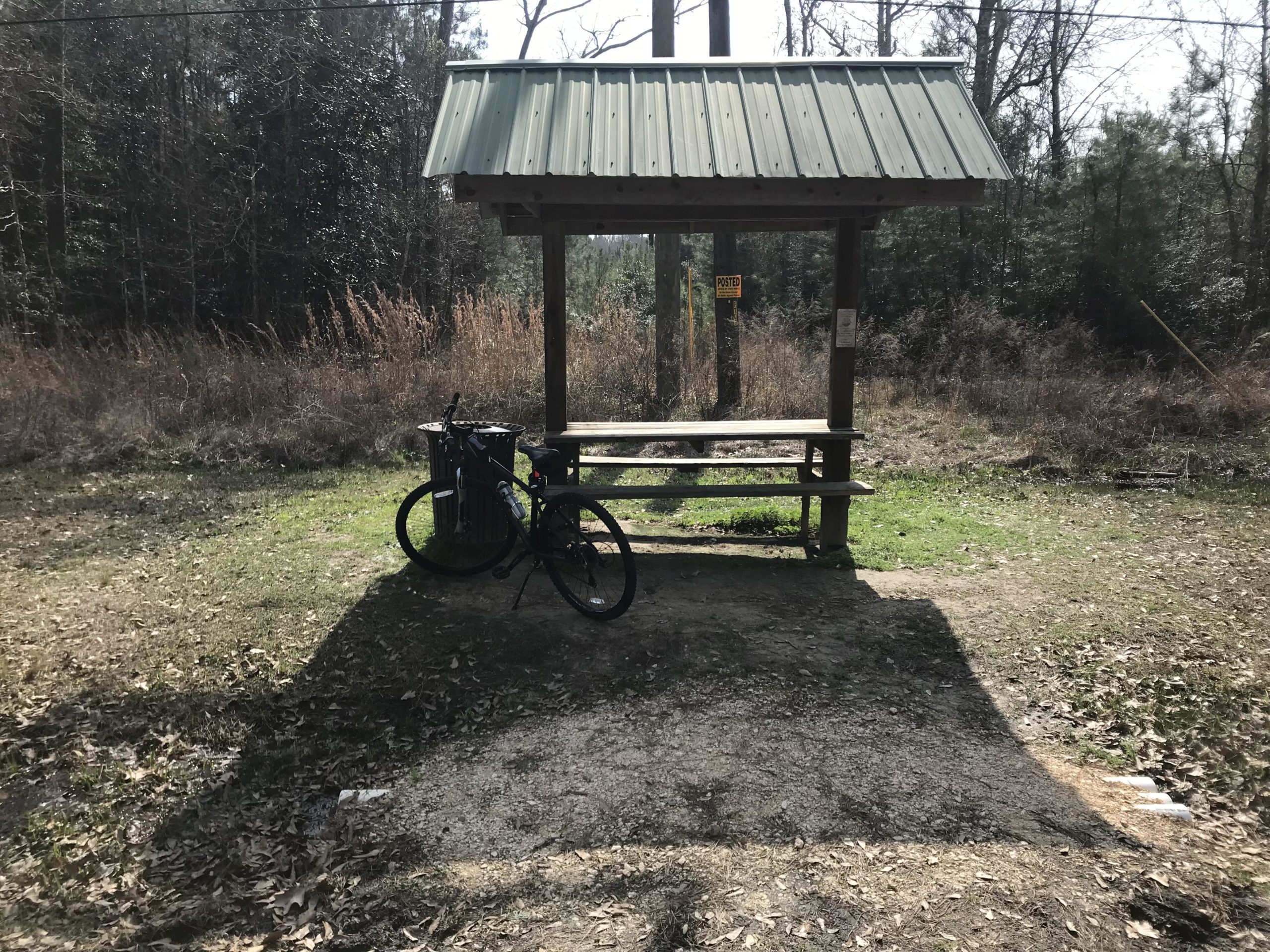 A bike parked beside a shelter with a wooden bench and metal roof, surrounded by tall grass and trees in a wooded area. A trash can is nearby, and a "posted" sign is visible in the background. The Tammany Trace mountain bike trail.