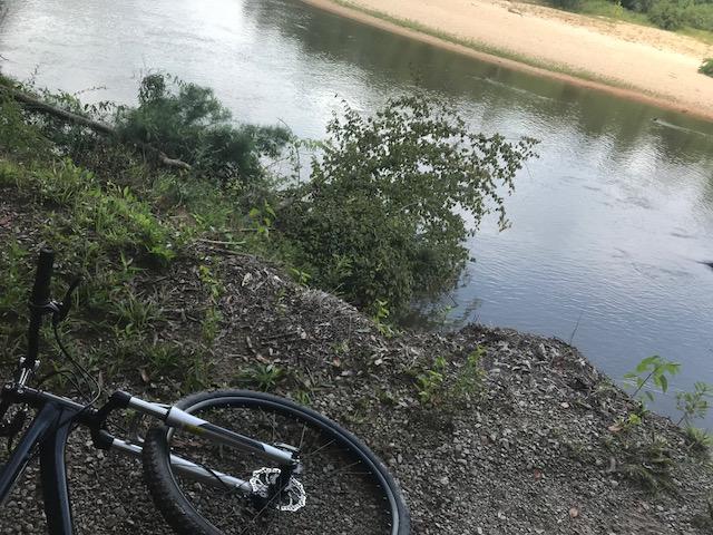 A bicycle rests on the ground near a riverbank, surrounded by green vegetation. The calm water of the river reflects the natural landscape, revealing a sandy area in the background. Bogue Chitto State Park mountain bike trail.