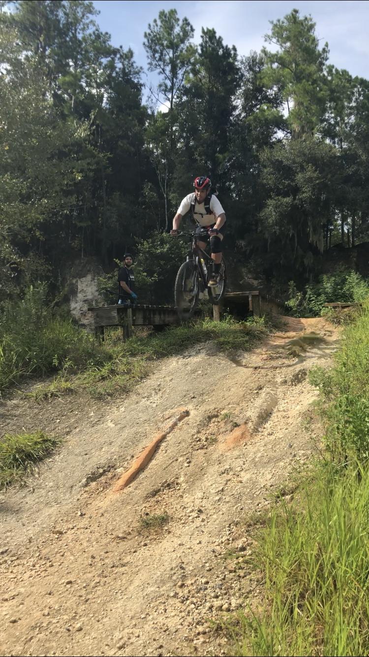 A mountain biker is captured mid-air as he jumps off a dirt ramp, with trees in the background. A person stands nearby, observing the action. The scene conveys a sense of adventure and excitement in an outdoor setting. Santos mountain bike trail.