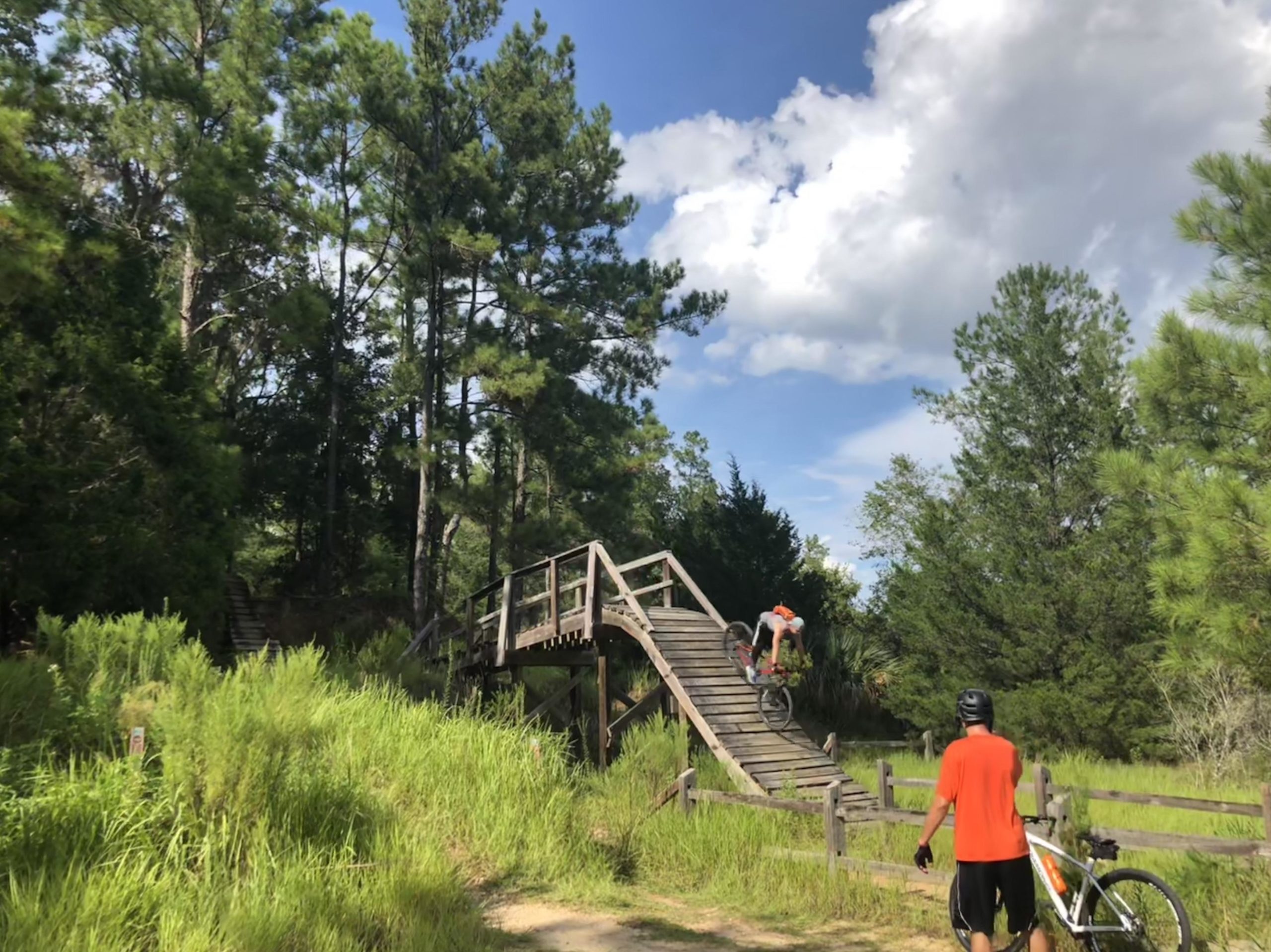 A mountain biker in mid-air jumps off a wooden ramp surrounded by tall trees and green grass, while another person in an orange shirt watches nearby with a bicycle. The sky is partly cloudy with blue peeking through. Santos mountain bike trail.