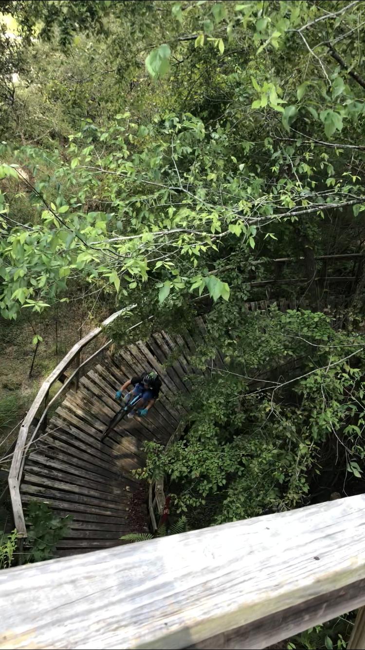 A bird's-eye view of a twisting wooden bike trail surrounded by lush green foliage, with a mountain biker navigating the curves. The image captures the natural beauty of the area and highlights the bike trail's construction. Santos mountain bike trail.