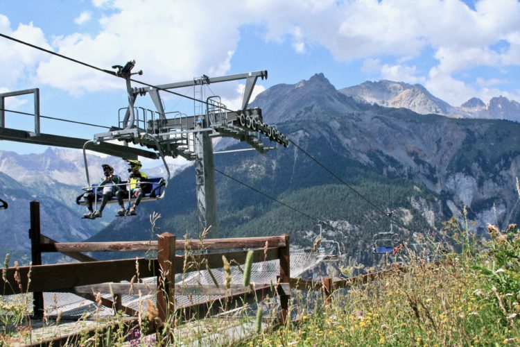 "Two people sitting on a chairlift, with mountain scenery and blue sky in the background. Wildflowers and a wooden fence are visible in the foreground."