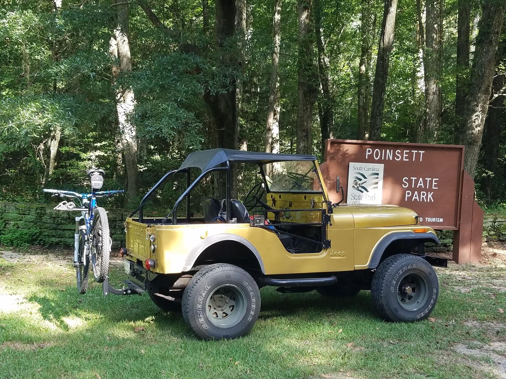 A vintage yellow Jeep parked next to a sign for Poinsett State Park, surrounded by lush green trees. A mountain bike is mounted on the back of the Jeep. The sign includes the South Carolina State Park logo. Poinsett State Park mountain bike trail.