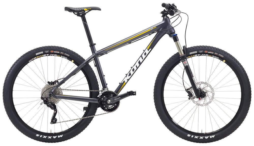 Kona Kona Kula: A side view of a modern mountain bike featuring a gray frame with yellow accents. The bike is equipped with wide tires, disc brakes, and a gear system suitable for off-road riding. The handlebars are straight, and the bike appears ready for trail adventures.