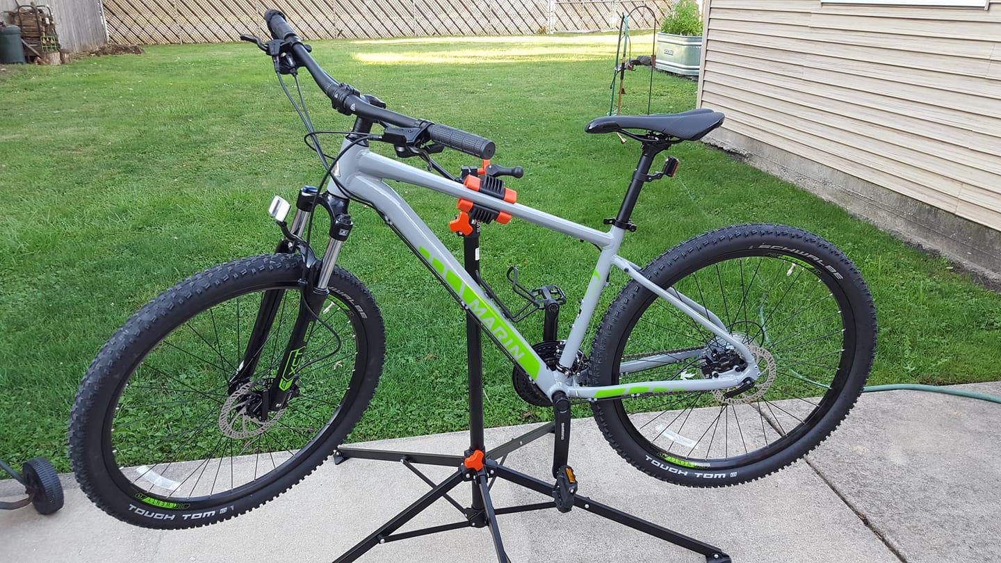 Marin Rock Spring 1: A gray and green mountain bike positioned on a repair stand in a grassy outdoor area. The bike features thick tires and front suspension, displaying its rugged design. The background includes a wooden fence and hints of a garden with tools.