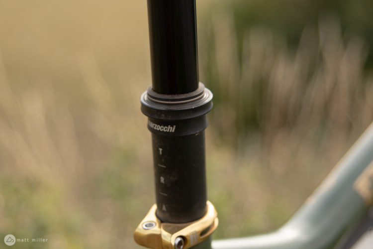 Close-up view of a bike's dropper seat post, featuring a black cylinder with visible measurement markings and a gold clamp at the base, set against a blurred natural background.