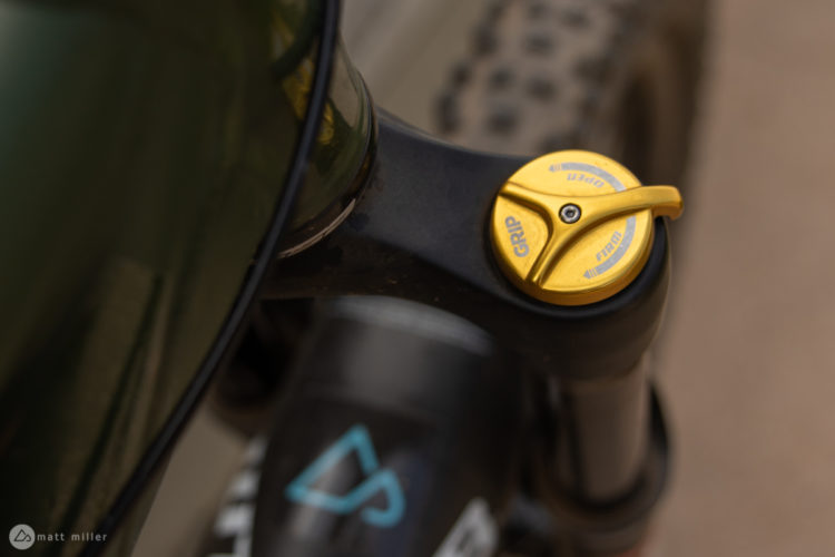 Close-up view of a mountain bike suspension adjustment knob, featuring a gold color. The knob is situated on a dark fork tube, with the word "GRIP" and various directional indicators visible. In the background, a portion of the bike's wheel and frame are partially visible.