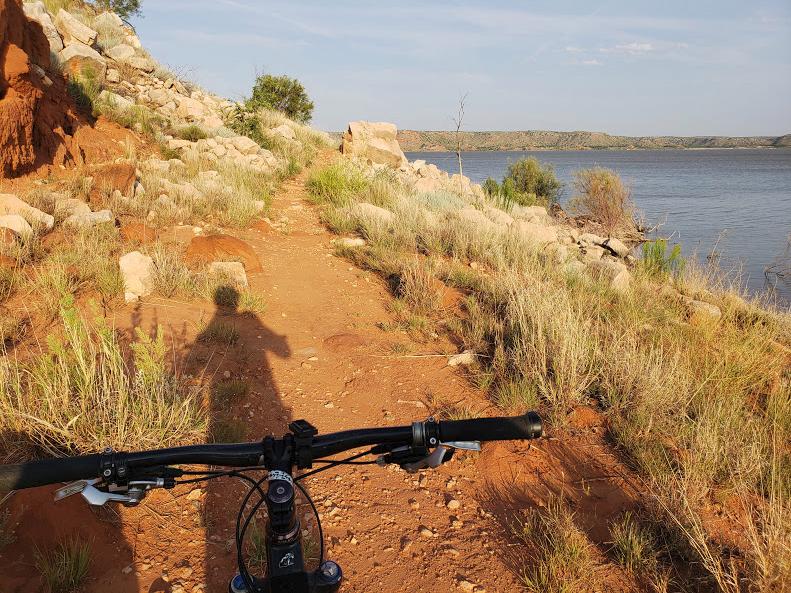 A mountain bike is positioned on a narrow dirt trail beside a body of water, surrounded by rocky terrain and sparse vegetation. The scene captures the natural landscape under a clear sky, with sunlight casting a shadow of the bike rider. Lake Meredith National Recreation Area mountain bike trail.