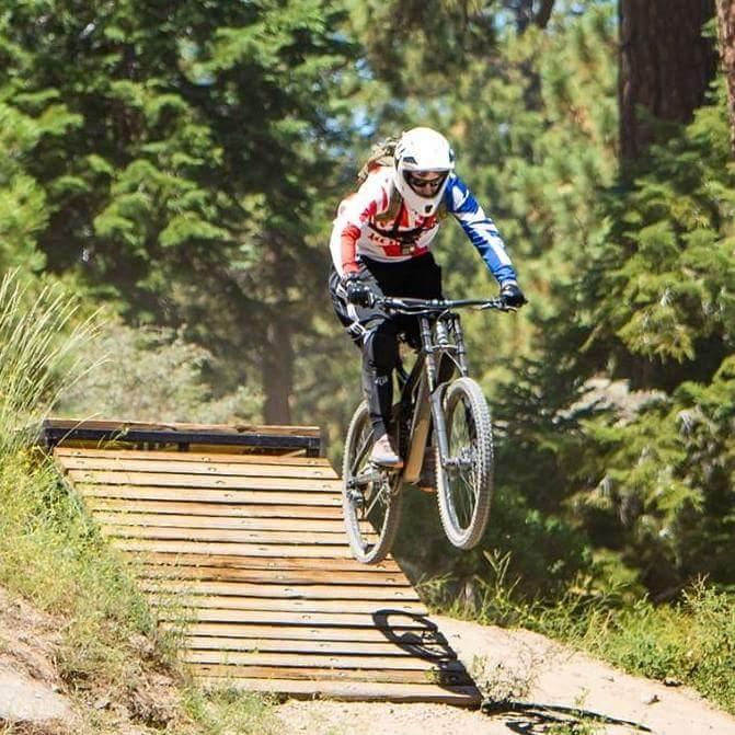 A mountain biker in a helmet and protective gear is mid-air as they jump off a wooden ramp surrounded by trees. The rider wears a red and blue jersey, showcasing a dynamic action shot against a natural background. Big Bear Mountain Resort mountain bike trail.