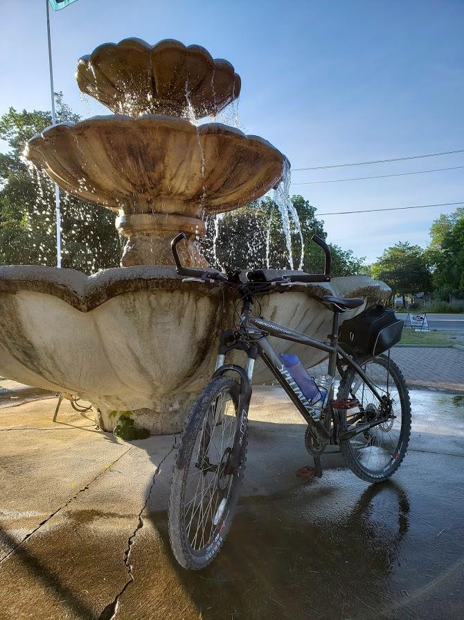 Specialized Hardrock Sport Disc: A bicycle parked near a stone fountain with multiple tiers, water flowing from the top tier. The scene is set in a sunny outdoor environment, with trees and a sidewalk visible in the background. The ground is wet, reflecting the sunlight, and a water bottle is attached to the bicycle.