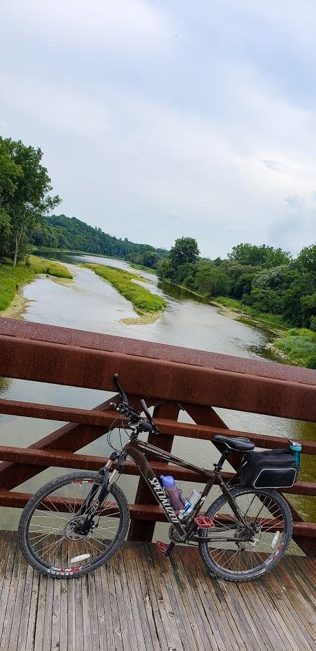 Specialized Hardrock Sport Disc: A mountain bike parked on a wooden bridge overlooking a calm river surrounded by lush greenery and rolling hills under a cloudy sky.