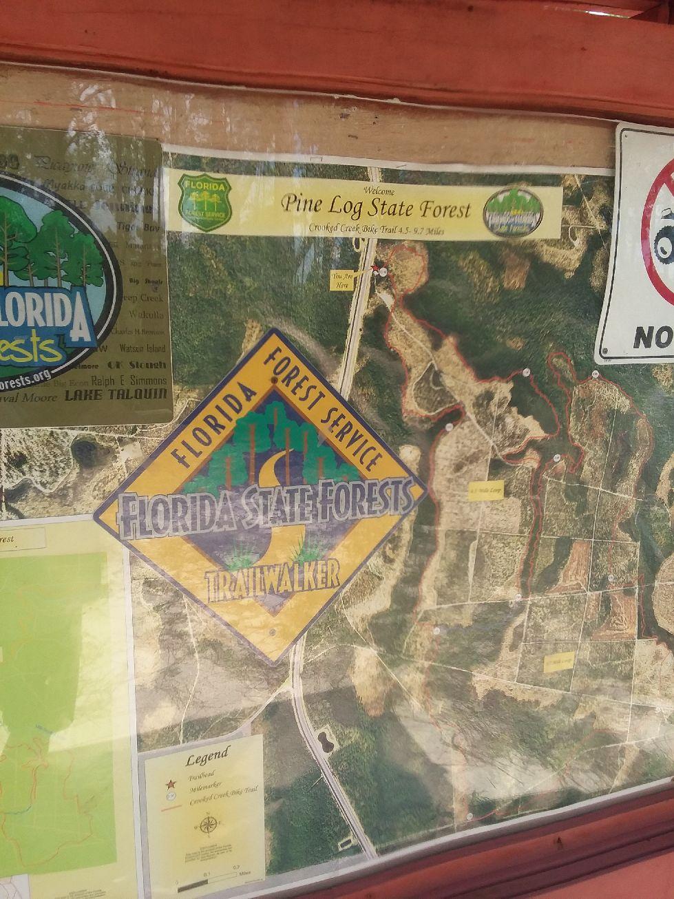 Map and informational sign for Pine Log State Forest, featuring trail details and a legend. The sign displays the Florida Forest Service logo and provides information about the Crawfish Creek biking trail, which is 4.5 to 9.7 miles long. Various annotations and logos related to Florida's state forests are also visible. Crooked Creek mountain bike trail.