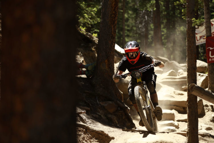 A mountain biker navigating a rocky trail through a forest, wearing a black shirt and red helmet. Dust is kicked up beneath the bike as the rider leans into the curve, surrounded by tall trees and a banner in the background.