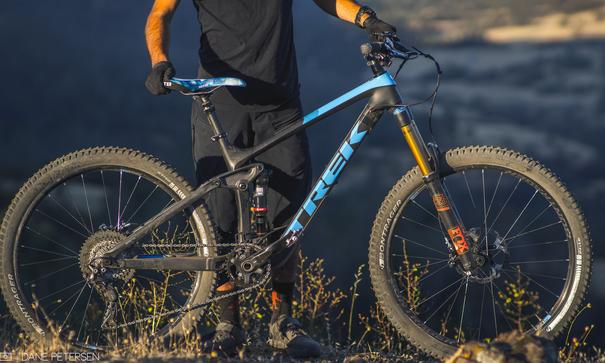 Trek Remedy 9.8 27.5/650b: A person stands on uneven terrain holding a blue and black Trek mountain bike. The bike features thick tires and a visible suspension system, indicating it's designed for off-road trails. The background shows a scenic landscape with hills and sunlight, suggesting an outdoor adventure. The individual is dressed in black athletic clothing and gloves, ready for biking.