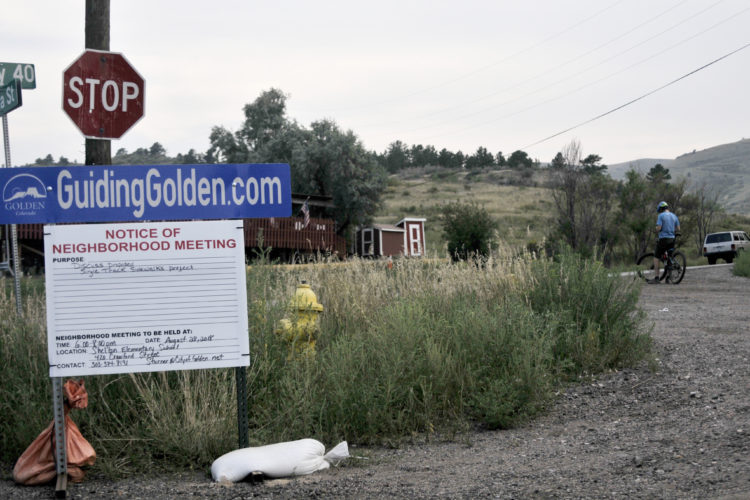 A stop sign and a notice of a neighborhood meeting are displayed on a post near a gravel road. The notice outlines details about discussing a proposed sidewalk project. In the background, a cyclist in a blue shirt and helmet is seen on the road, while a yellow fire hydrant and small buildings are visible amidst the grassy landscape. The image reflects a community-focused environment in a rural setting.