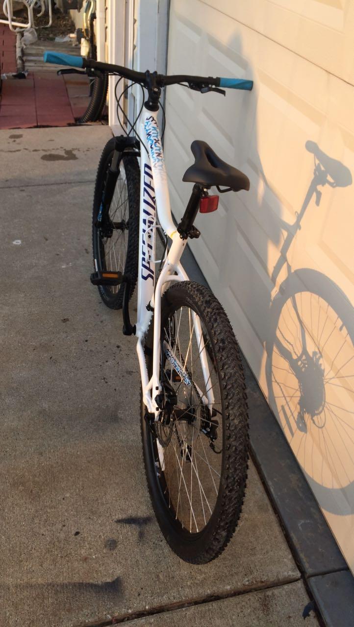 Specialized Specialized jinx: A white mountain bike with blue accents is parked on a concrete surface, leaning against a garage door. The bike features thick, knobby tires and a saddle positioned for comfortable riding. The sun casts a shadow of the bike on the ground, highlighting its shape and details.