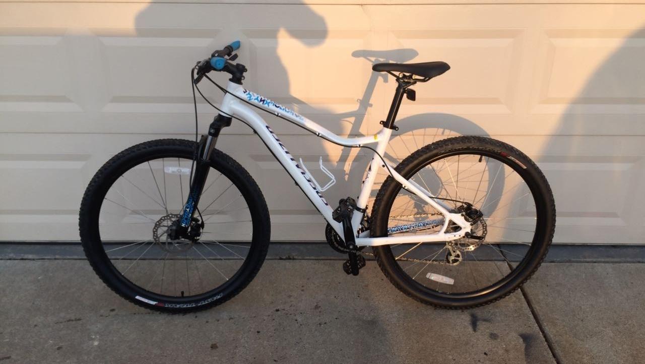 Specialized Specialized jinx: A white mountain bike with black tires and components, leaning against a beige garage door. The bike features a front suspension fork, blue handlebar grips, and gear shifters. The photo captures the bike in a well-lit setting, emphasizing its design and features.