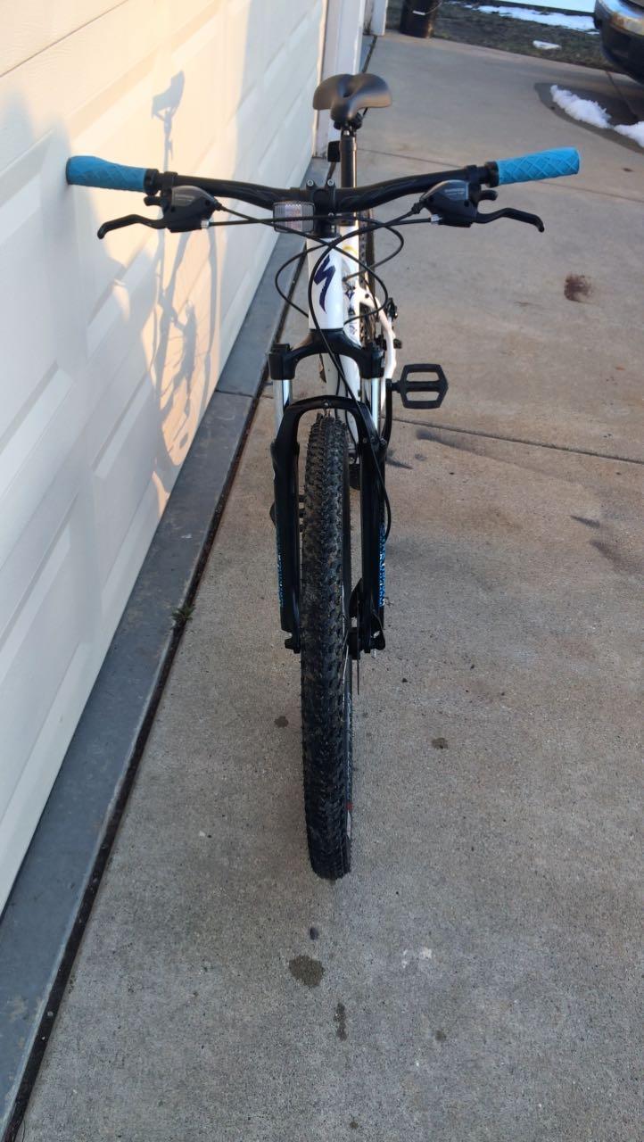 Specialized Specialized jinx: A white and black mountain bike viewed from the front, showing the handlebar with blue grips and the front wheel on a concrete surface. In the background, a garage door is visible, along with some patches of snow on the ground.