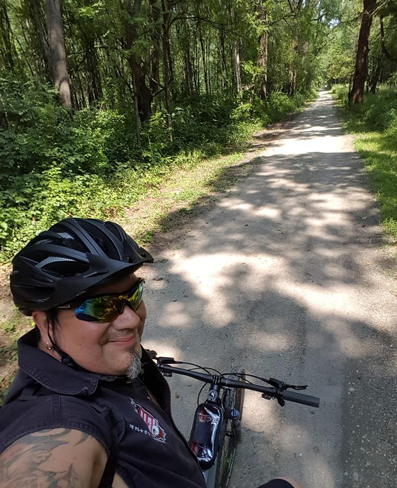 A person wearing a black helmet and sunglasses is sitting on a mountain bike on a dirt path surrounded by lush green trees. The sunlight filters through the foliage, casting dappled shadows on the trail. The individual appears to be enjoying a leisurely ride in a natural setting. Palos Forest Preserve mountain bike trail.