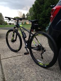 Trek Xcaliber 9: A black and green mountain bike parked on a concrete driveway, with a parked car and a residential area visible in the background.