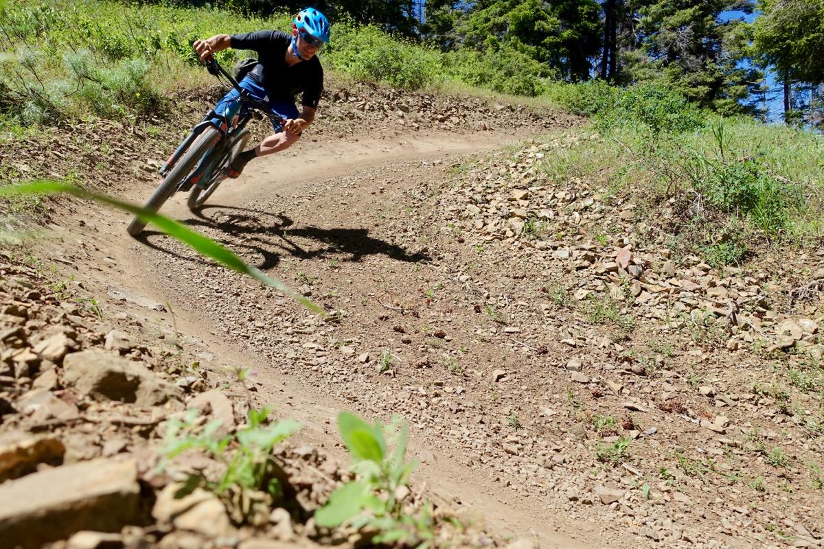 A mountain biker wearing a blue helmet leans into a corner on a dirt trail surrounded by greenery. The cyclist is captured mid-action, showcasing a dynamic riding posture as they navigate a curving path lined with rocks and plants. Mind Bender mountain bike trail.