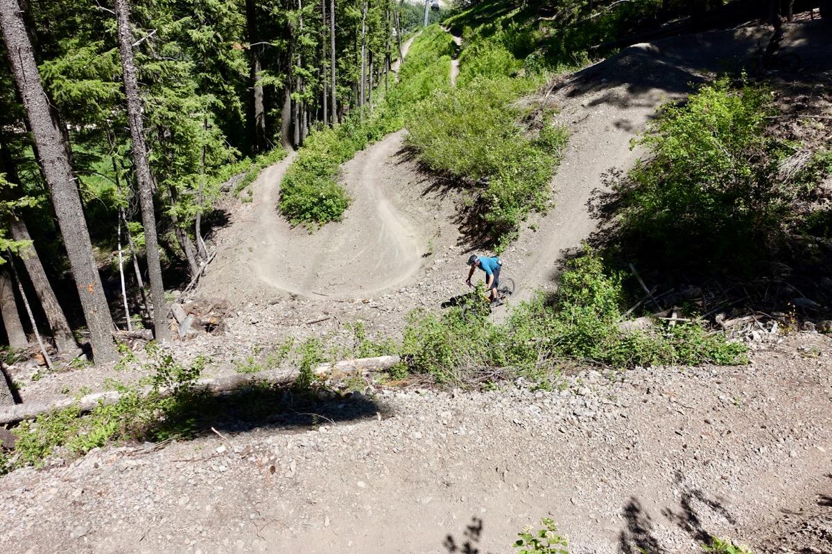 A mountain biker riding on a dirt trail surrounded by tall trees, with winding paths leading through a green forest. The terrain is rocky and uneven, indicating an adventurous mountain biking route. Pale Rider mountain bike trail.
