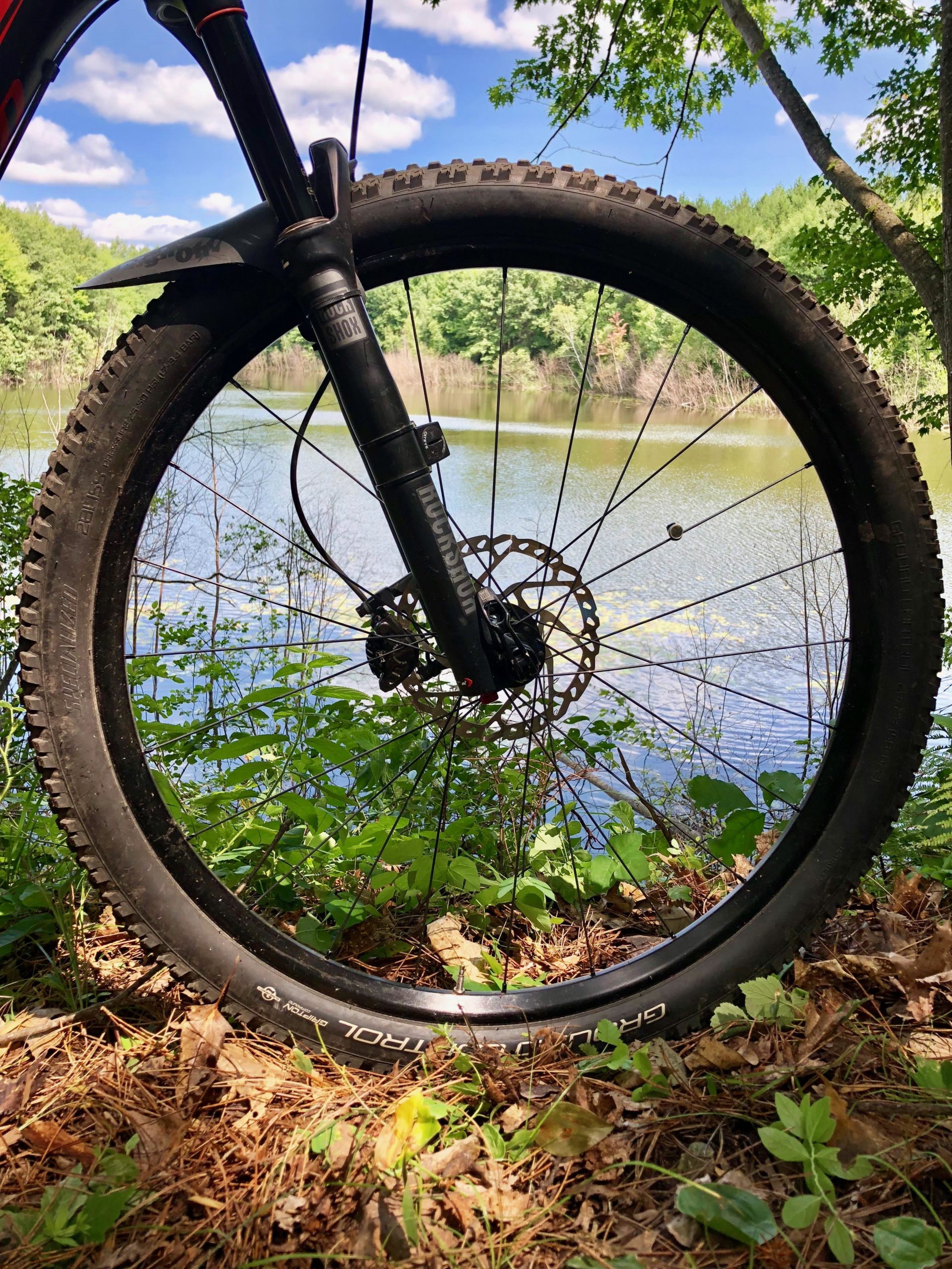 Close-up view of a mountain bike wheel, situated near a serene lake surrounded by greenery. The image captures the tire tread and fork of the bike against a backdrop of water and trees, under a bright blue sky with fluffy clouds. Hickory Ridge mountain bike trail.