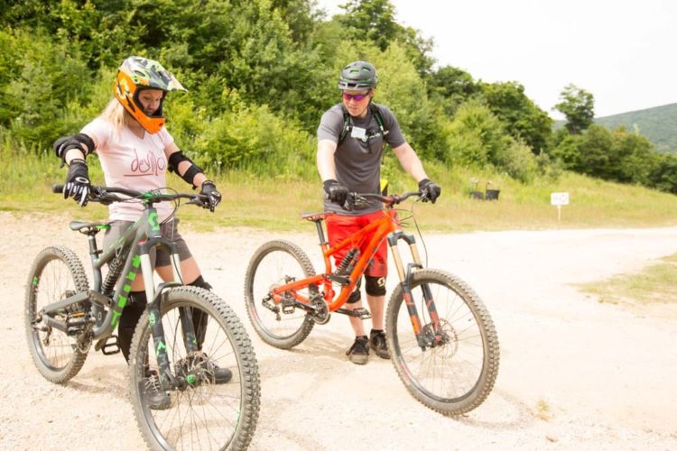 A young girl in a helmet and protective gear stands next to a bicycle, listening attentively to an adult man who is pointing at her bike. Both are on a gravel path surrounded by greenery, indicating an outdoor setting for mountain biking. The man is wearing a helmet and casual biking attire, emphasizing a coaching or mentoring moment.
