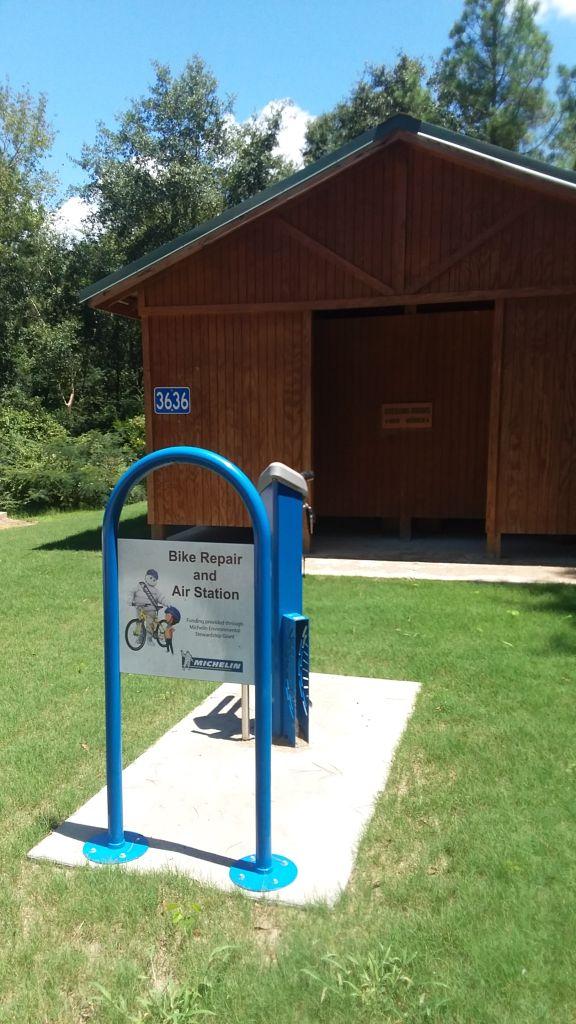 Blue bike repair and air station in front of a wooden building, with a sign that reads "Bike Repair and Air Station." Surrounded by green grass and trees under a clear sky. The building has the number 3636 displayed on it. Beaver Creek / Forever Wild Trails mountain bike trail.