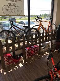 A bicycle store interior showcasing two bicycles mounted on a wooden fence display by a window, with several boxes underneath. The view outside shows parked cars and a sunny day.