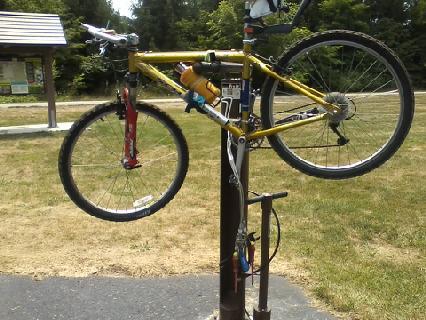 A yellow mountain bike mounted on a vertical bike repair stand in a grassy park area. The foreground shows the bicycle hanging securely, while a picnic shelter and trees are visible in the background. Oak Openings -- Beach Ridge Singletrack Trail mountain bike trail.