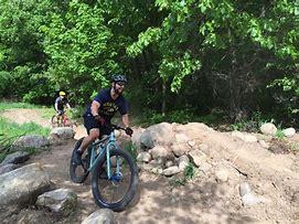 A person riding a mountain bike on a rocky trail in a lush, green outdoor setting. Another cyclist is visible in the background, also navigating the trail. DTE Energy Foundation Trail mountain bike trail.