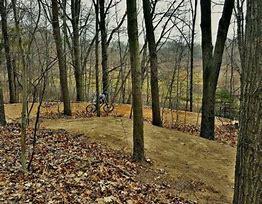 A cyclist riding on a dirt trail through a wooded area, surrounded by leafless trees and fallen leaves on the ground, with a view of a distant landscape in the background. DTE Energy Foundation Trail mountain bike trail.
