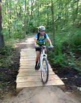 A child riding a bicycle on a wooden bridge in a forested area, surrounded by greenery and trees. The child is wearing a helmet and a blue shirt, enjoying an outdoor biking adventure. DTE Energy Foundation Trail mountain bike trail.