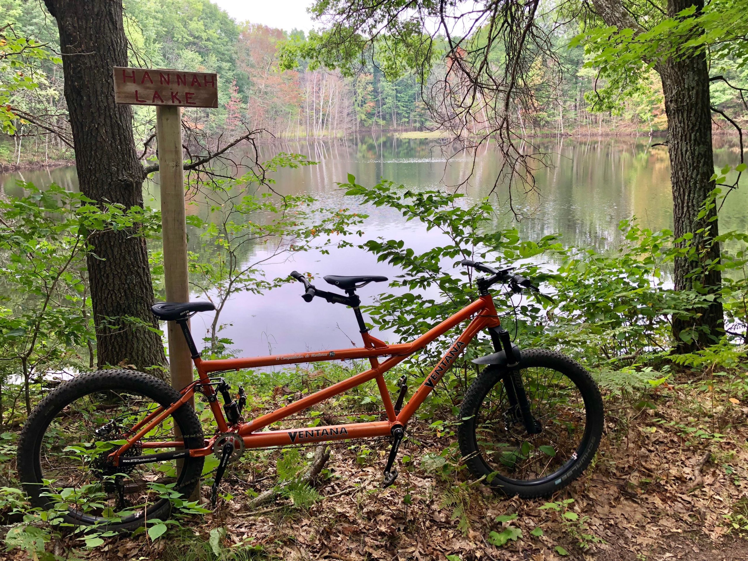 A scenic view of Hannah Lake, featuring a wooden sign with the lake's name. In the foreground, an orange mountain bike is leaning against a tree, surrounded by lush greenery and leaves, with the calm water of the lake reflecting the surrounding trees in the background. Hickory Ridge mountain bike trail.