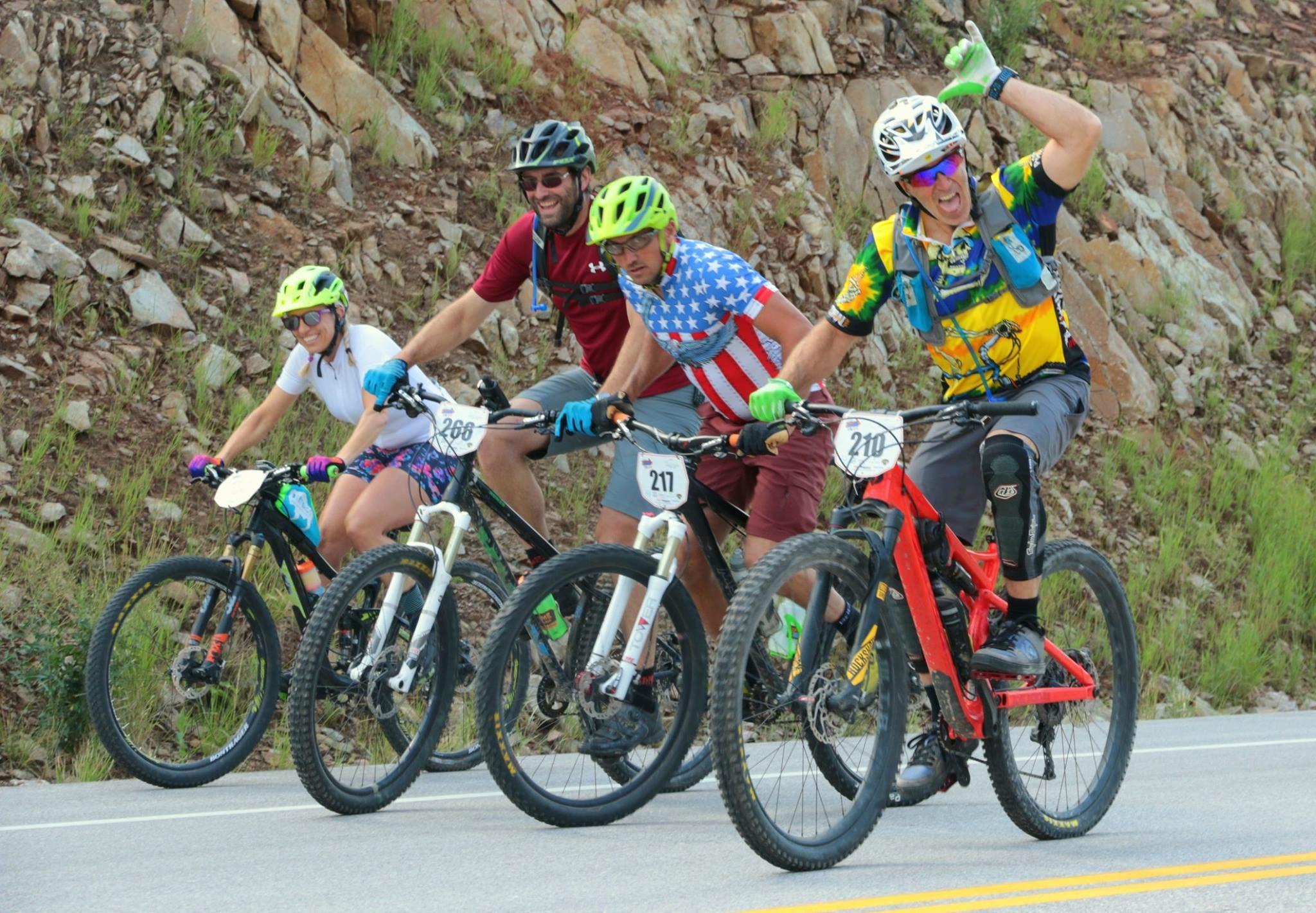 Salsa Deadwood Sus: A group of four mountain bikers, all wearing helmets and colorful outfits, pose together on a rural road. Two women and two men, one of whom is excitedly raising his hand in a gesture of triumph, are positioned on their bikes, showcasing vibrant bike models. The background features rocky terrain and patches of grass, indicating an outdoor cycling event. Each biker displays a race number on their bike.