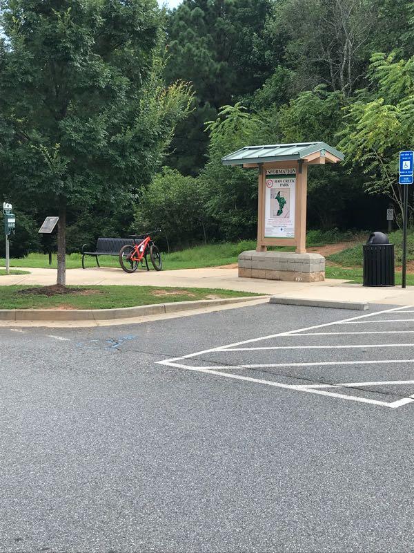 A scenic outdoor area featuring a bike leaning against a bench near a trailhead. There is a sign displaying a map of the park's trails, surrounded by trees and green foliage. A nearby trash can and handicap-accessible parking space are visible in the parking lot. Haw Creek Park mountain bike trail.