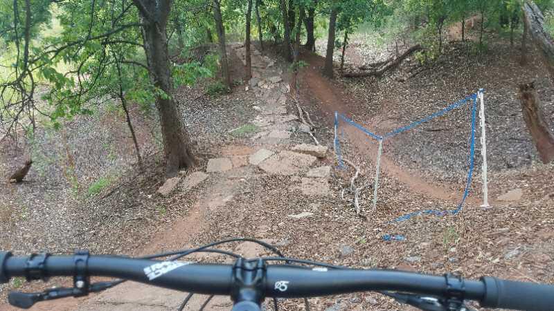 A rocky bike trail viewed from the handlebars of a mountain bike, surrounded by trees and greenery. The path features natural stone steps and a section with a wooden barrier on the right, indicating a winding trail through a forested area. Wee-chi-ta mountain bike trail.