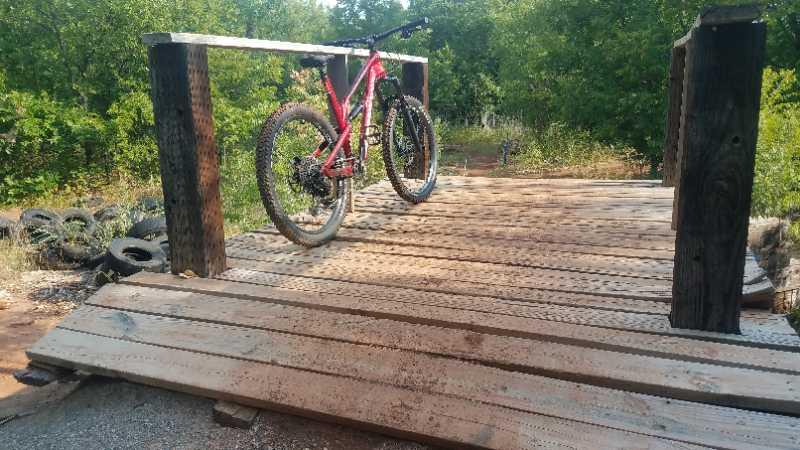 A pink mountain bike resting on a wooden ramp that leads into a natural setting with trees in the background. The ramp is constructed of planks and held up by sturdy wooden posts, with some tires visible in the foreground. The scene suggests an outdoor biking or trail area. Wee-chi-ta mountain bike trail.