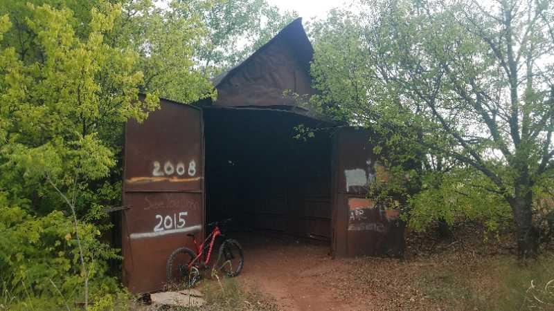 A rusted metal shed partially hidden by trees, with a bicycle parked next to it. The shed has graffiti markings indicating the years "2008" and "2015" on its doors. Wee-chi-ta mountain bike trail.