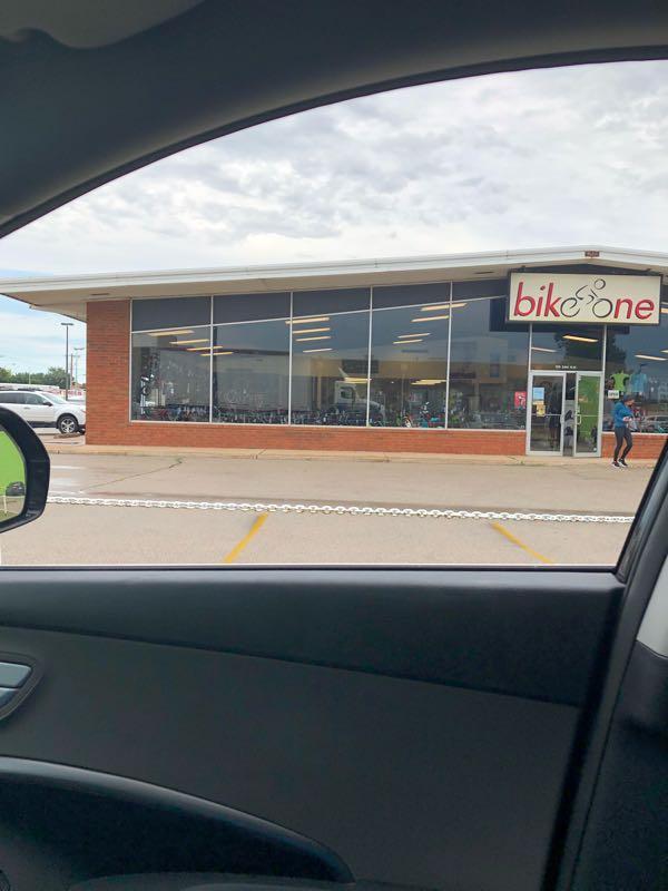 Image of a bicycle shop named "bike one," featuring large glass windows showcasing various bicycles inside. The storefront is brick, and a person is standing outside near the entrance. The image is taken from inside a vehicle, showing a portion of the car door and side mirror. The sky appears cloudy.