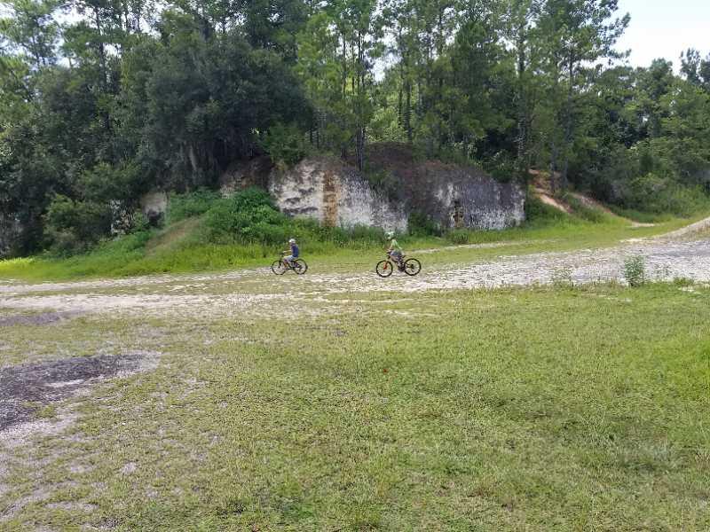 Two children riding bicycles on a gravel path surrounded by green grass and trees. In the background, there are rocky hills partially covered in vegetation. The scene is set in a natural outdoor environment. Santos mountain bike trail.